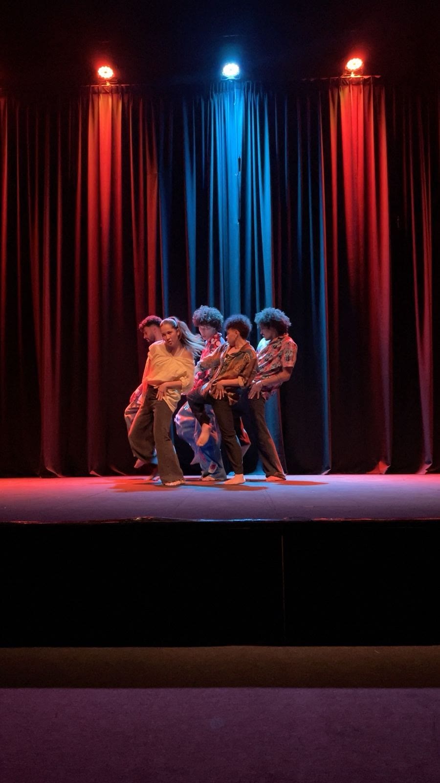 Five dancers press together in a line on a stage lit red and blue
