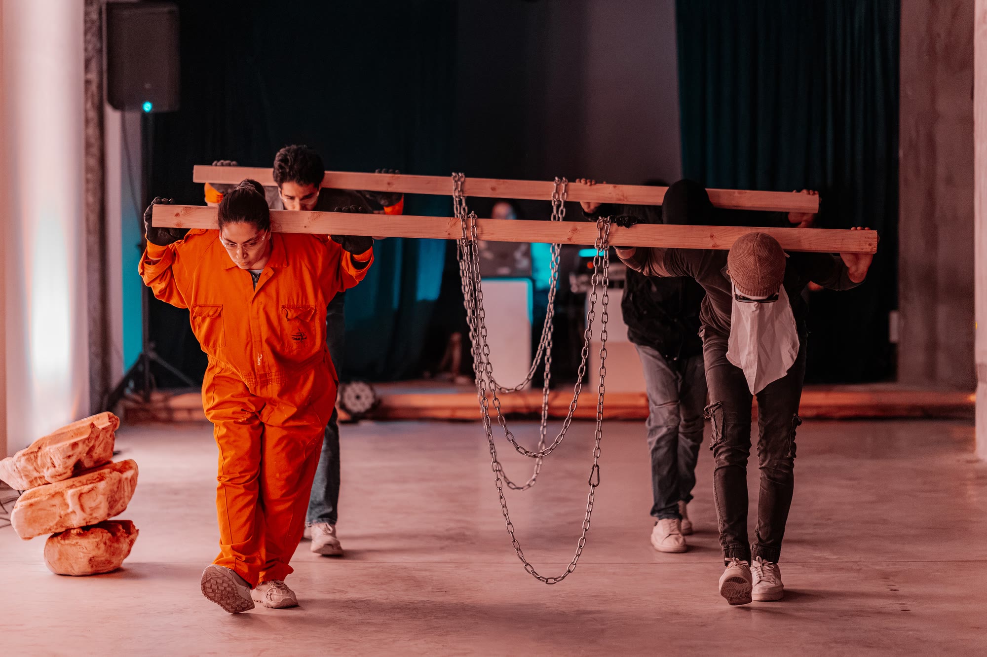 Performers carry timber beams hung with chains across their shoulders; one wears an orange jumpsuit