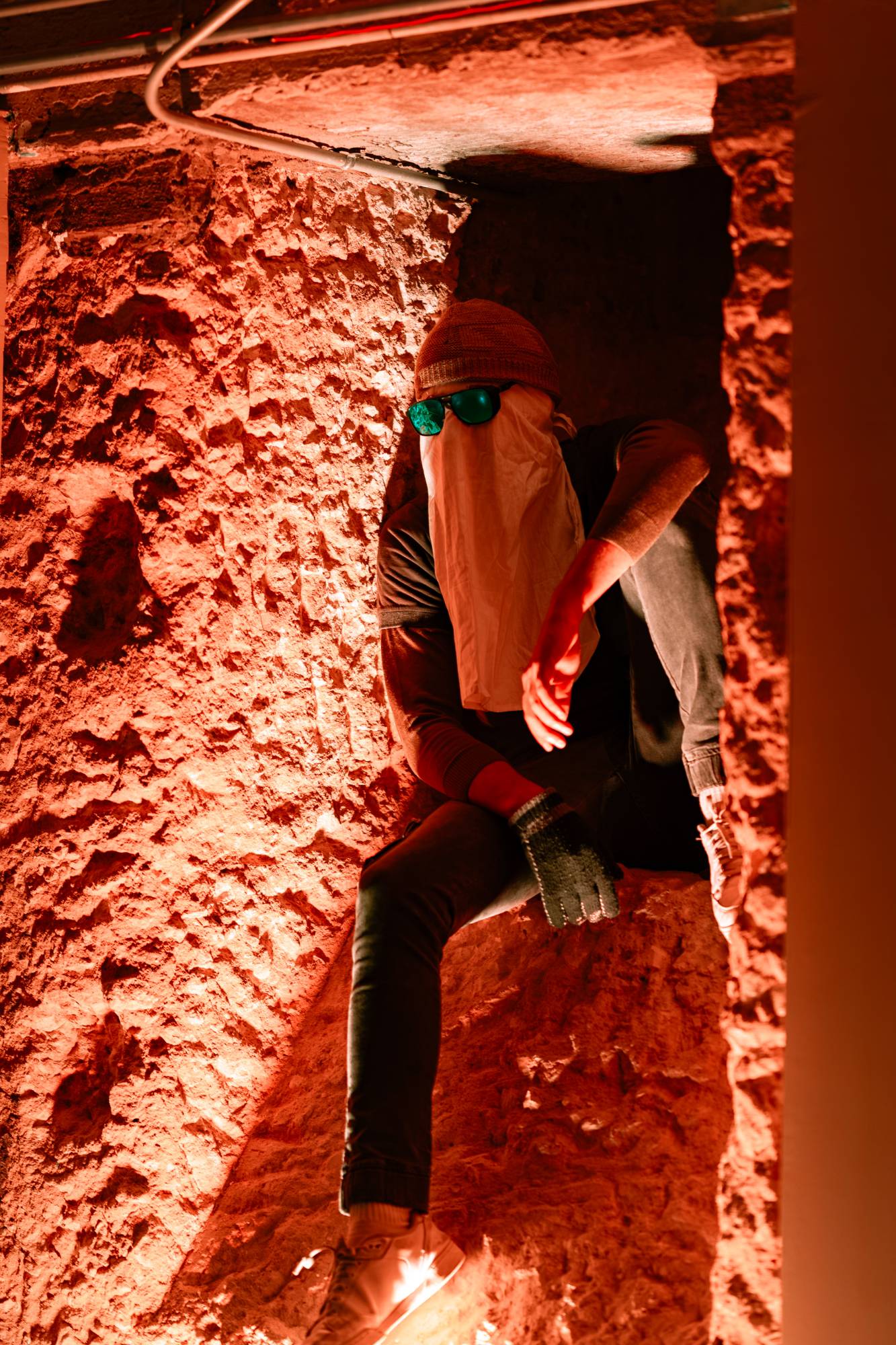 A performer with a cloth over his face and green goggles sits wedged in a red-lit stone recess