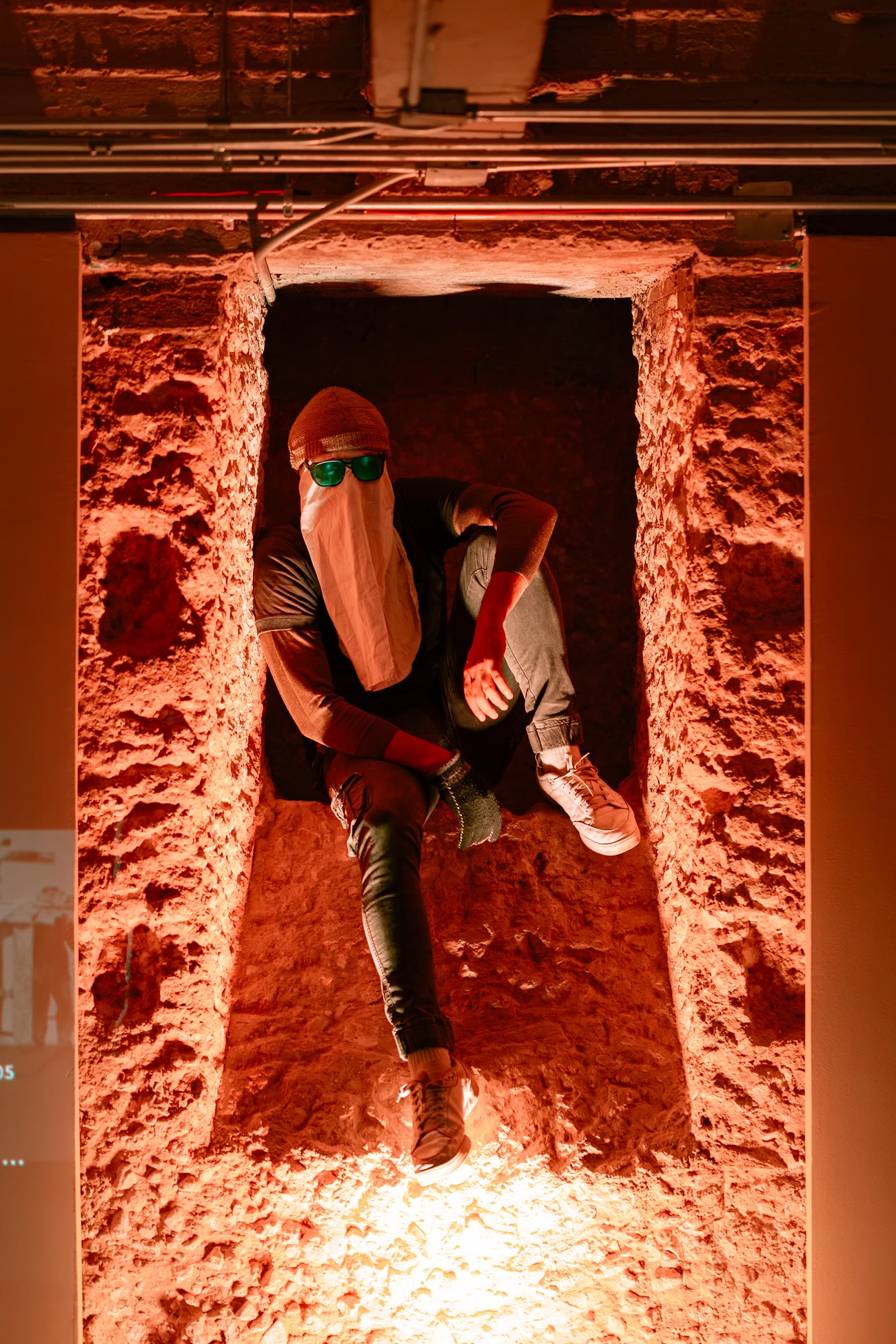 A masked performer in green goggles crouches inside a red-lit stone niche