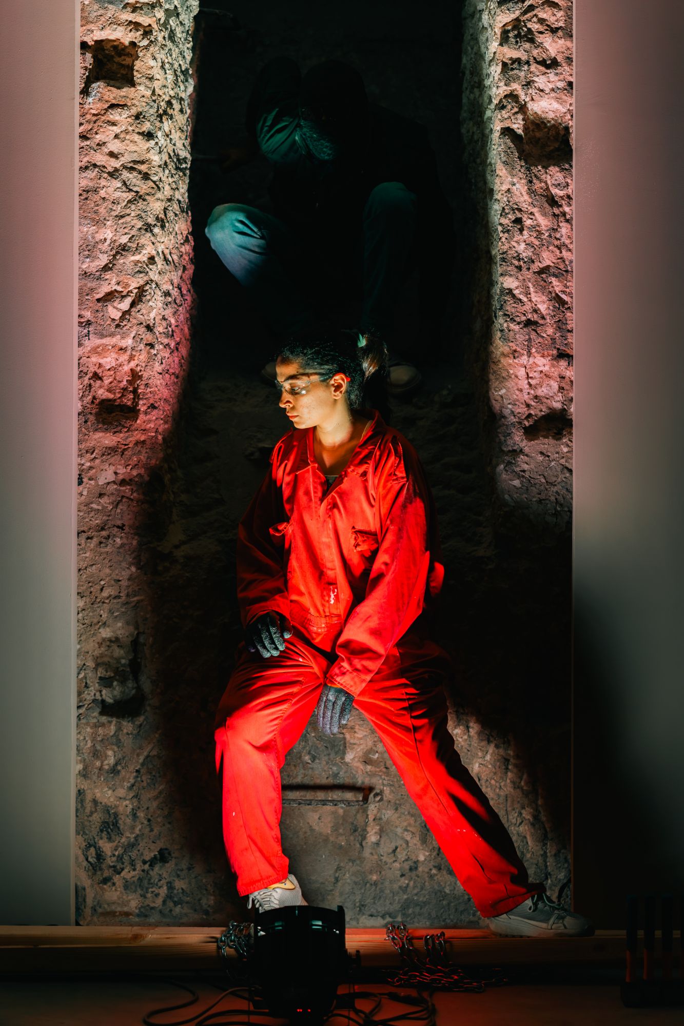 A performer in a red jumpsuit and safety glasses sits in a stone niche