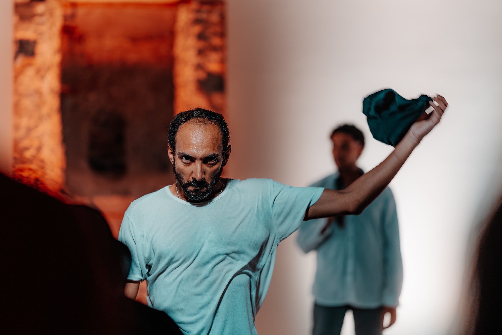 A performer in a white T-shirt holds his balaclava at arm's length and looks straight at the camera